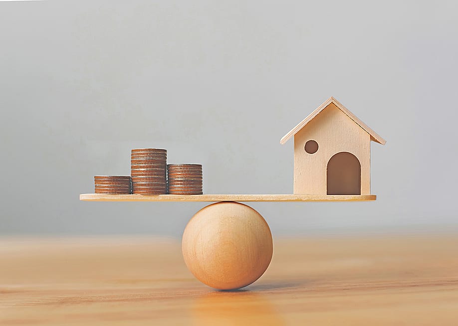 Wooden scale balancing a mini wooden figure of a house on the right and stack of coins on the left. — stefanzisus // Shutterstock