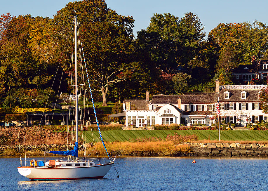 A sailboat moored off-shore near a luxury waterfront home in Greenwich, Connecticut. — James Kirkikis // Shutterstock