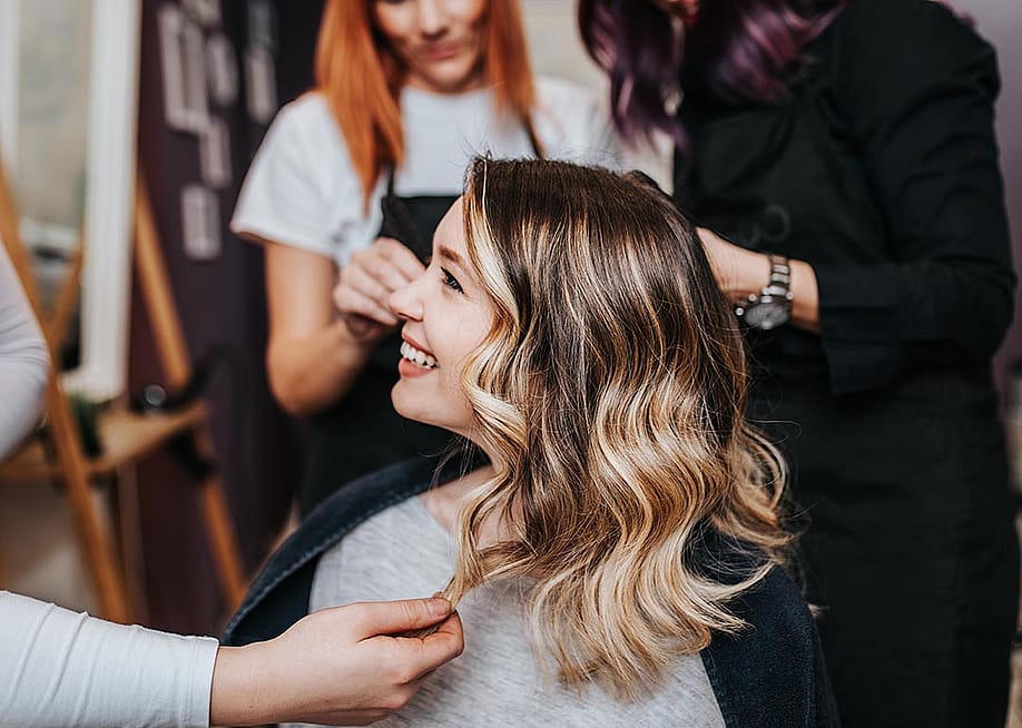 Side profile of woman with blond and brunette hair in long layers smiling at hair salon with two stylists behind her in the background. — hedgehog94 // Shutterstock