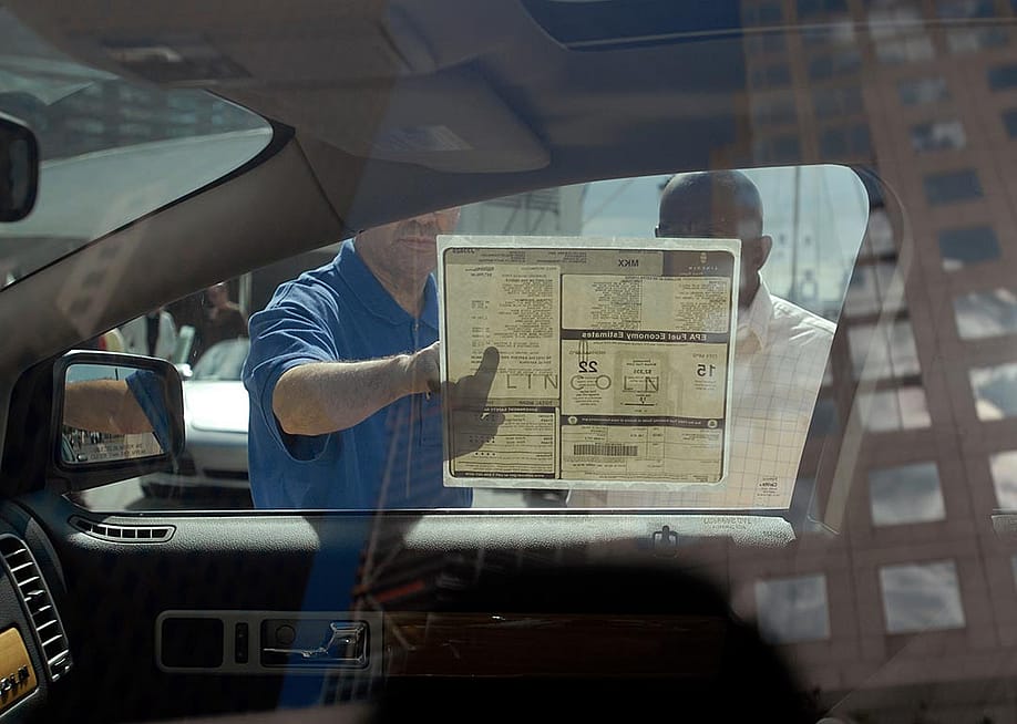 Interior view of car from driver's side looking out passenger window where two people are reading a sticker on the window with the car's features. — rblfmr // Shutterstock