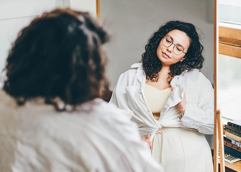 A curvy woman stands in front of a mirror, styling her outfit with a long sleeve shirt. — Mariia Korneeva // Shutterstock