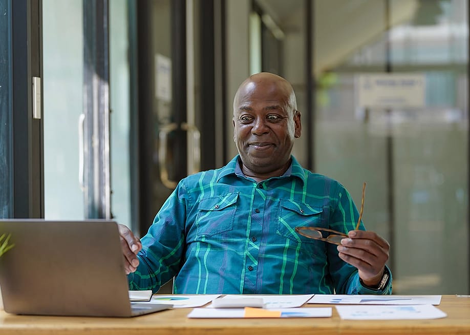 A senior man excitingly looks at financial documents while using a laptop. — Songsak C // Shutterstock