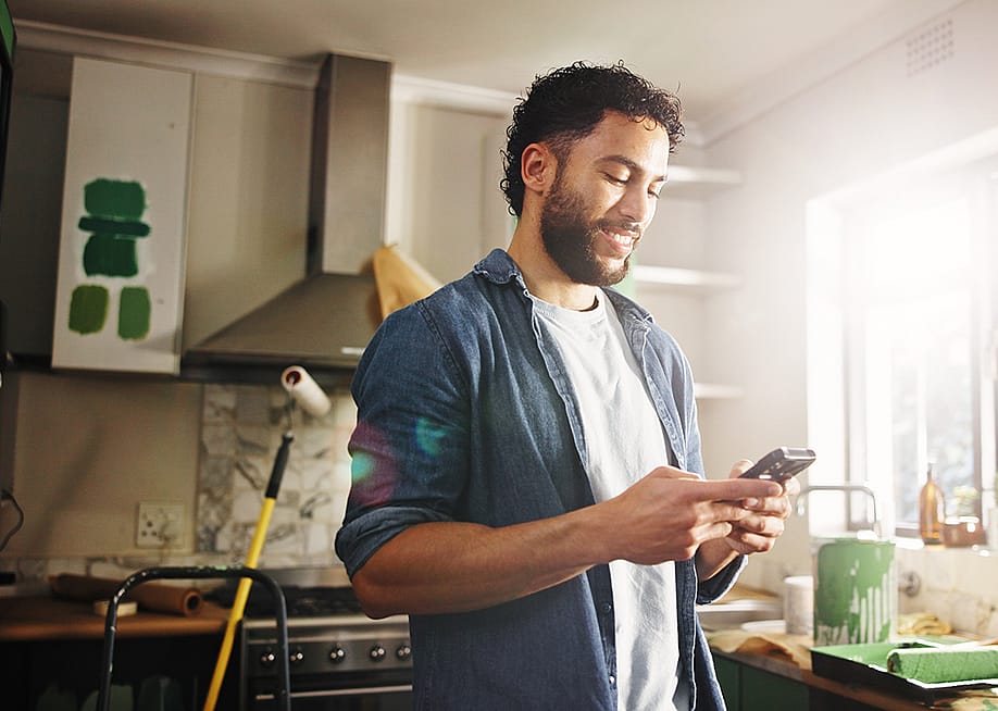 A man his apartment's kitchen looking up renovation ideas in his phone. — PeopleImages // Shutterstock