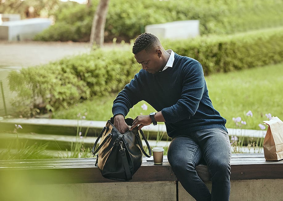 A young black businessman, looking inside his bag to eat lunch while sitting on a bench at a park. — PeopleImages // Shutterstock