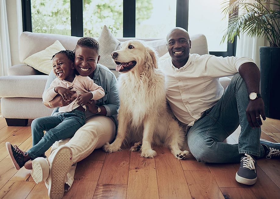 A black family of three and their dog, happily sitting on the living room floor of their home. — PeopleImages // Shutterstock