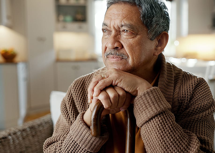 A senior man with a cane thinks deeply while sitting on a sofa. — PeopleImages // Shutterstock