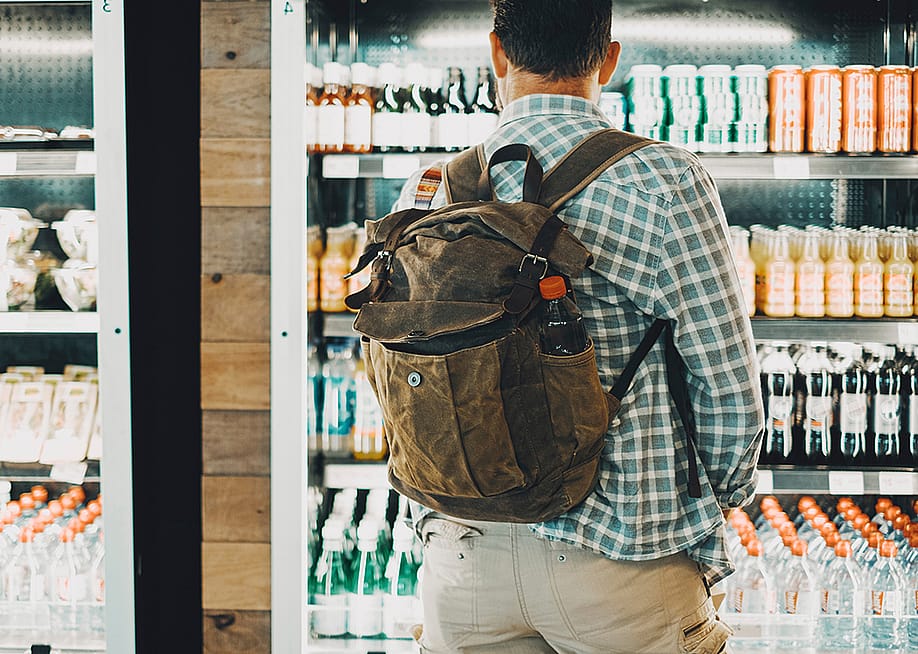 A man traveling with a backpack facing the cold drinks section of an airport food store. — simona pilolla 2 // Shutterstock