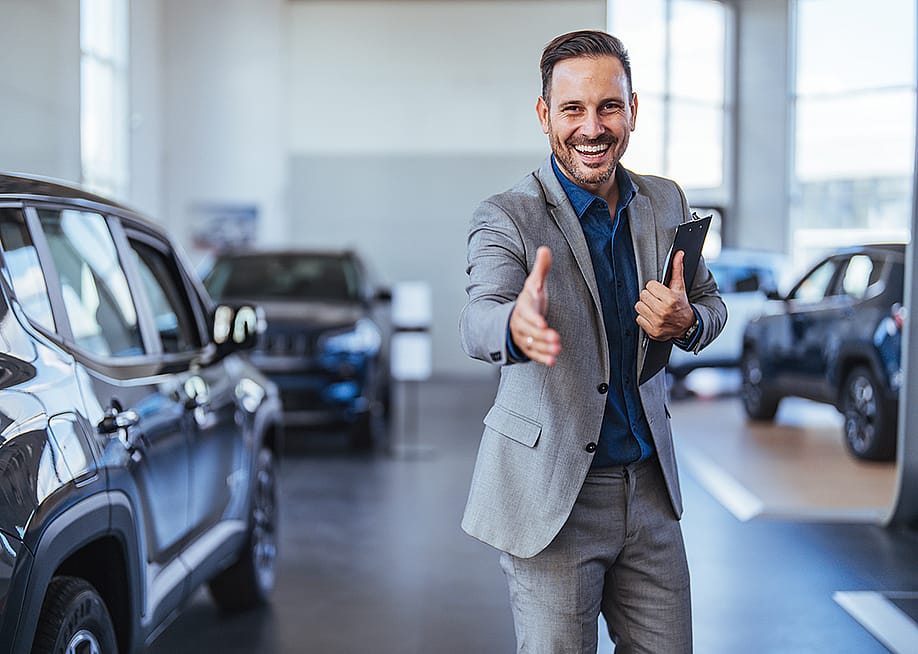 A friendly and cheerful salesman poses to greet car showroom clients with a handshake. — Dragana Gordic // Shutterstock