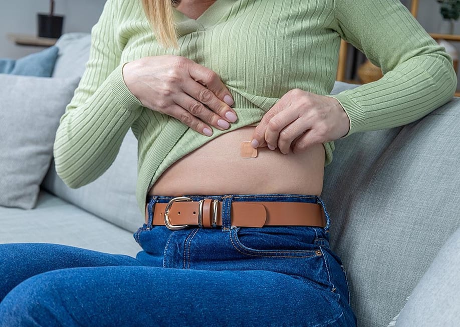 Close up of woman sitting on couch pulling up long sleeve light green shirt to apply hormone replacement patch to her abdomen. — Dmytro Zinkevych // Shutterstock