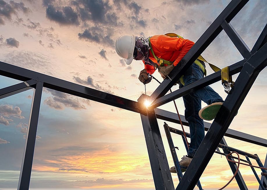 Low angle view of a welder is welding steel on a steel roof truss with the sunrise in the background. — Tong_stocker // Shutterstock