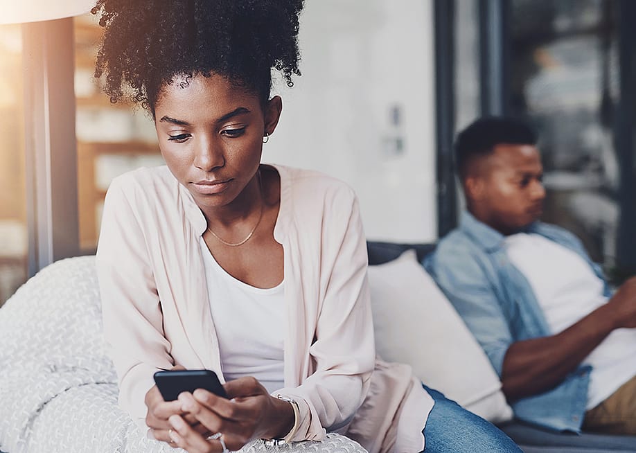 A black couple on their individual phones while sitting apart on the same couch at home. — PeopleImages // Shutterstock