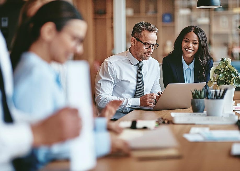 Two diverse businesspeople smiling while working on a laptop together at the end of a boardroom table in an office. — Ground Picture // Shutterstock