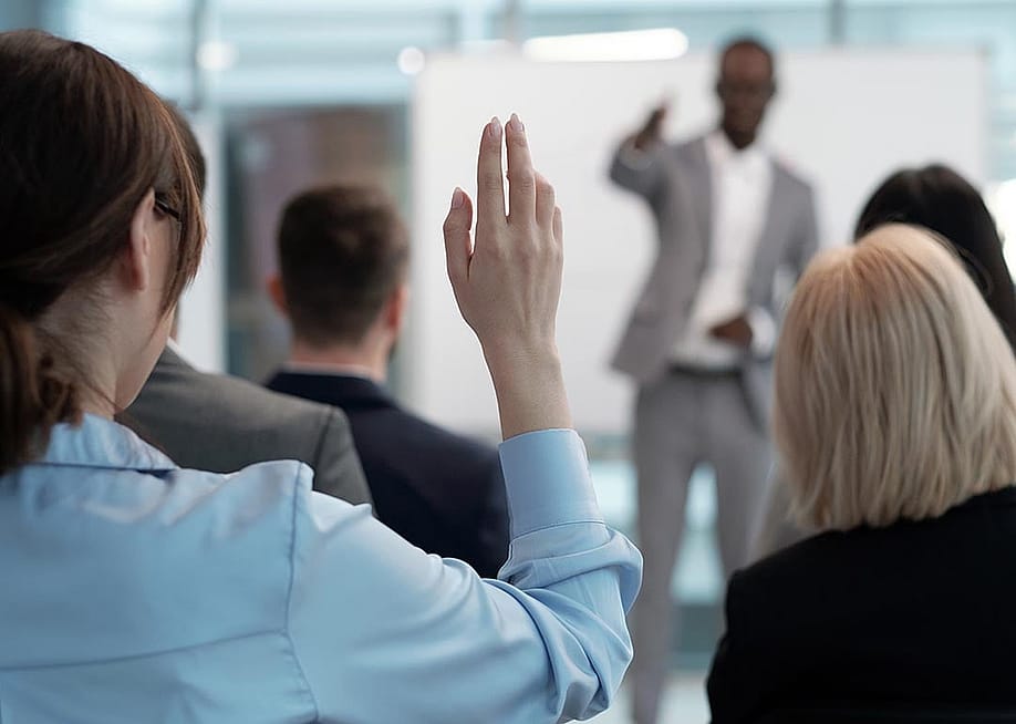 Rear view of raised hand with pointy nails of young employee wishing to answer question of lecturer while sitting behind group of other speakers at conference. — Pressmaster // Shutterstock