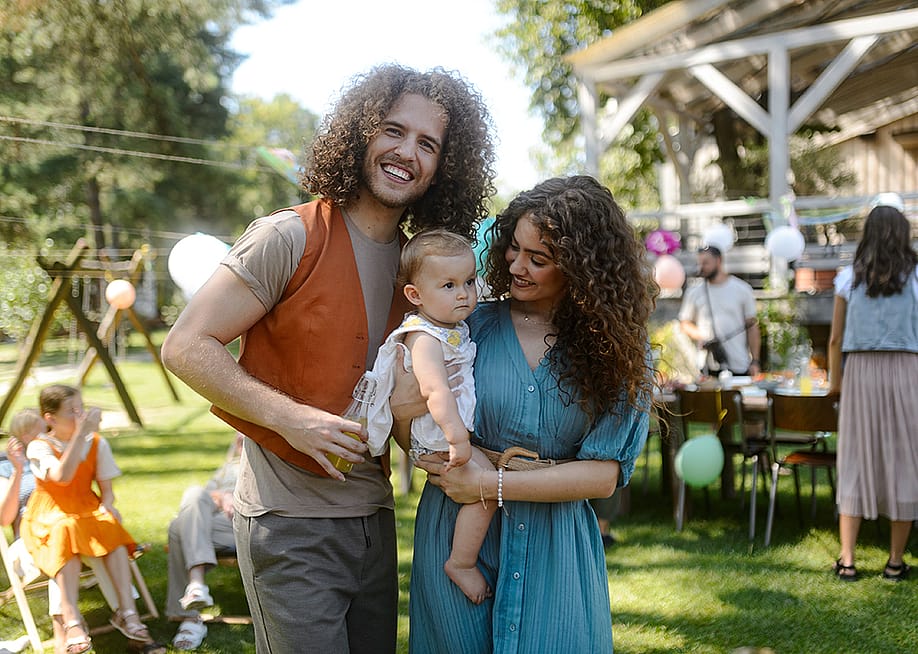 A happy couple pose with their baby at a backyard party. — Halfpoint // Shutterstock