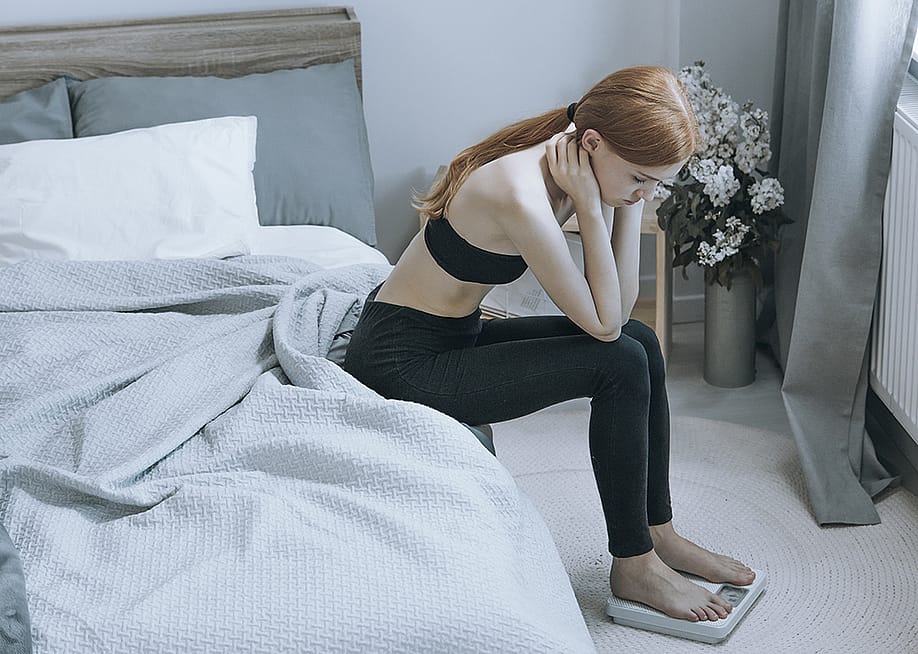 A young woman sits by her bed while pensively checking weight on a scale. — Ground Picture // Shutterstock