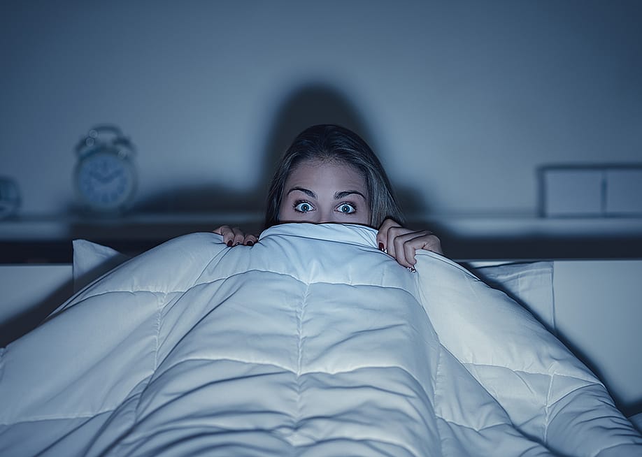 Stylized photo of woman hiding under covers with wide eyes, illuminated by screen from TV. — Stock-Asso // Shutterstock