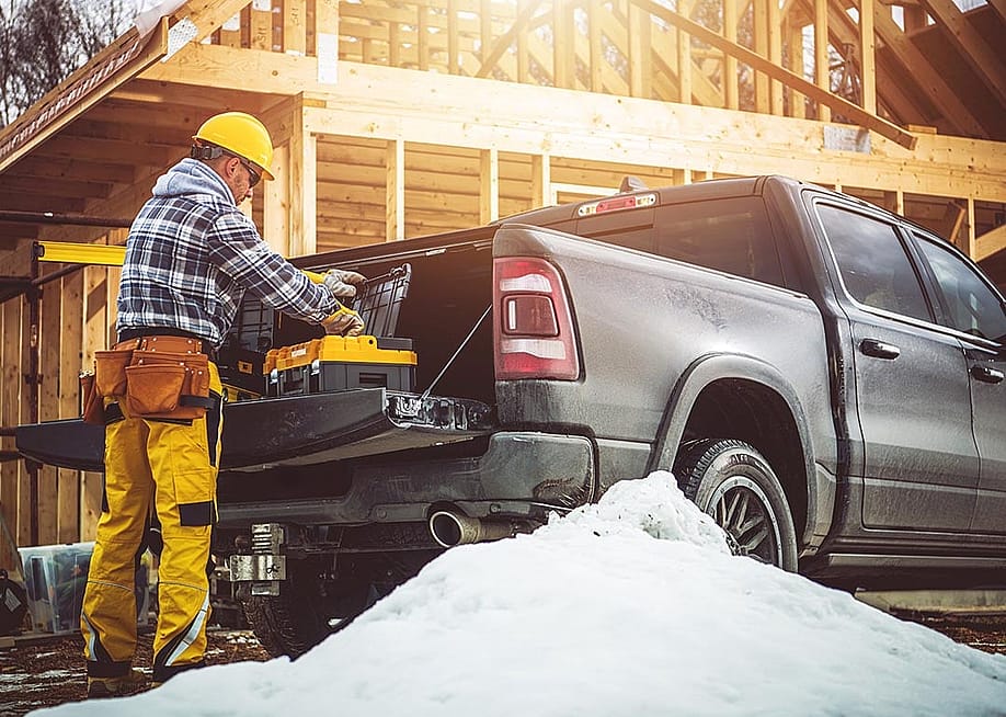 A construction worker in yellow gear prepares tools from a truck at a snowy building site with wooden building framing in the background. — Virrage Images // Shutterstock.