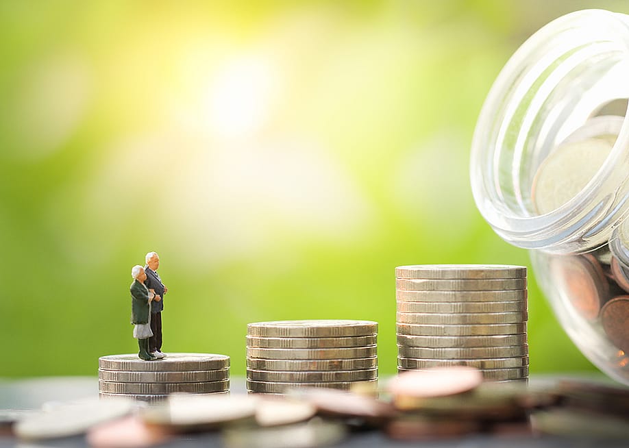 A miniature elderly couple stands on a stack of coins leading to a glass jar full of savings. — Behopeful // Shutterstock