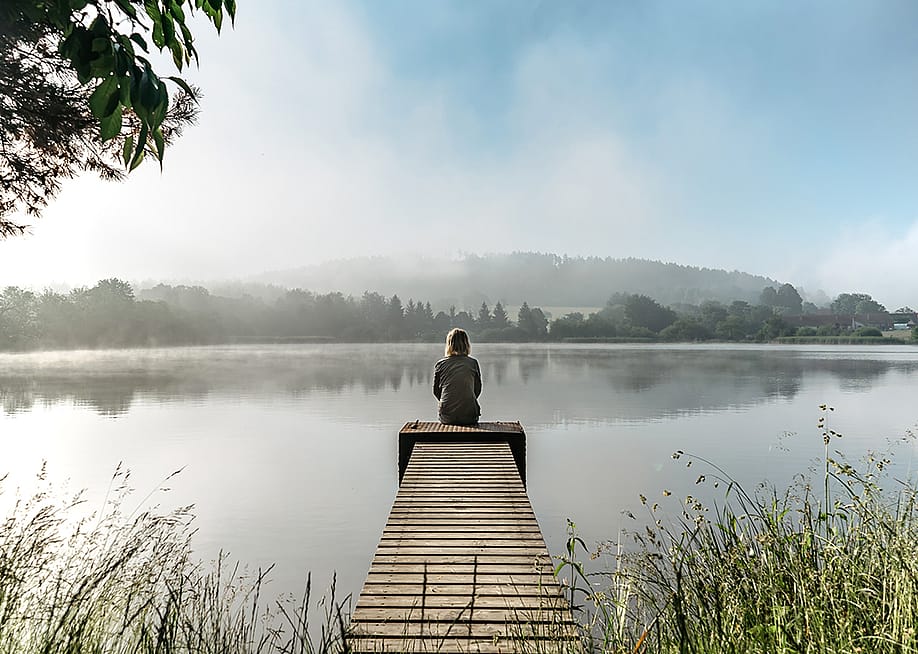 A woman sits quietly at the end of a small pier by a lake during a calm, misty morning. — Eva Pruchova // Shutterstock