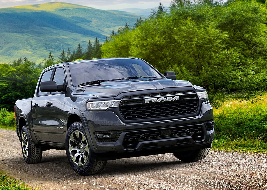 Black 2026 Ram 1500 REV Bighorn driving up dirt road with rolling green hills in background. — Stellantis