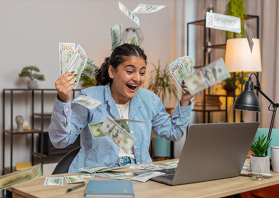 Young person throwing money and bills in the air in front of a computer; concept of getting rich quickly myth — Andrii Iemelianenko // Shutterstock