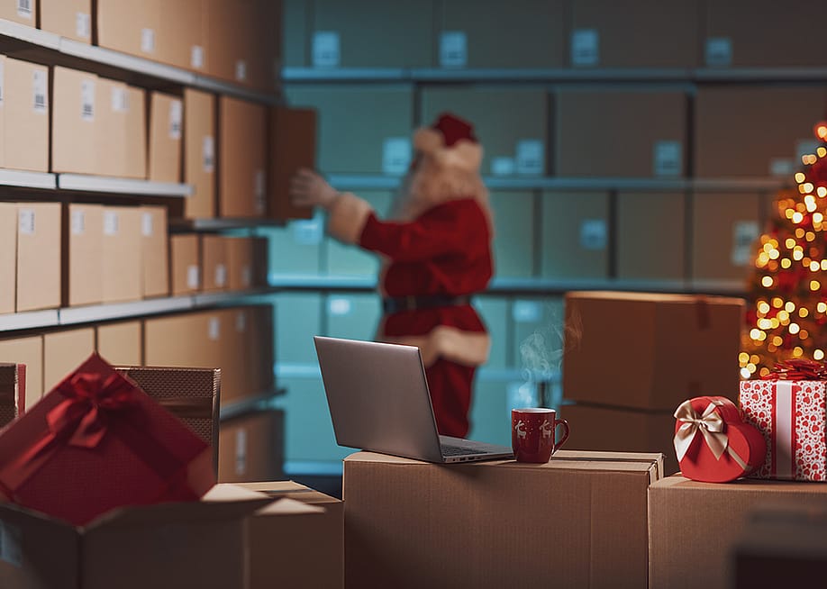 A worker dressed as Santa Claus prepares delivery Christmas gift boxes in a warehouse. — Stock-Asso // Shutterstock