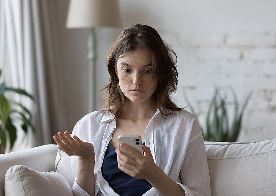 A woman sitting on a couch at home looking concerned at her phone. — fizkes // Shutterstock