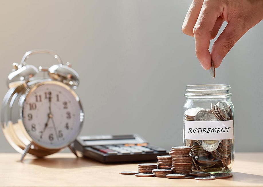 Hand putting coins in glass jar labeled "retirement" next to calculator and analog alarm clock with window in above in the background. — chayanuphol // Shutterstock