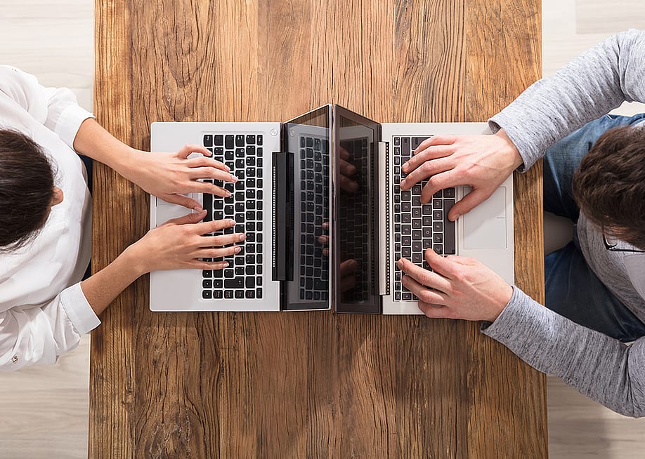 An overhead view of two people using laptops and sitting across each other on a table. — Andrey_Popov // Shutterstock