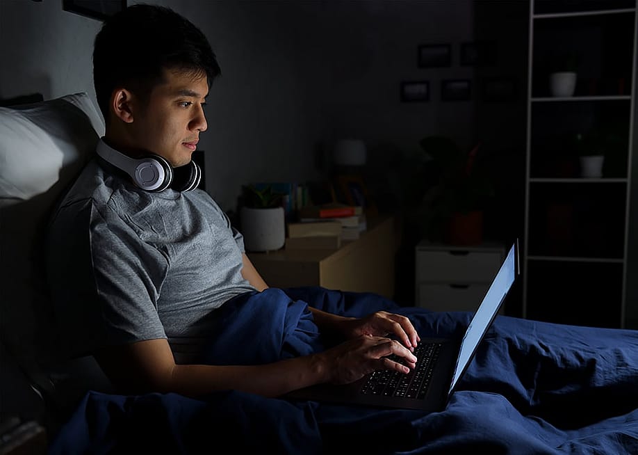 A young man uses his laptop in bed at night. — amenic181 // Shutterstock