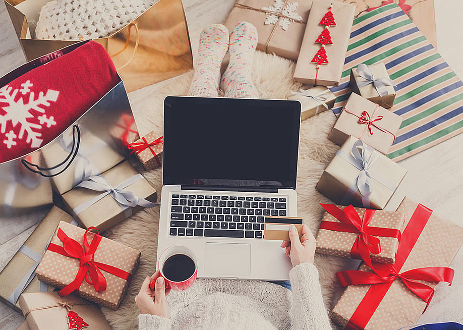 A shopper uses a laptop and credit card while sitting on a bed surrounded by Christmas gifts. — Prostock-studio // Shutterstock