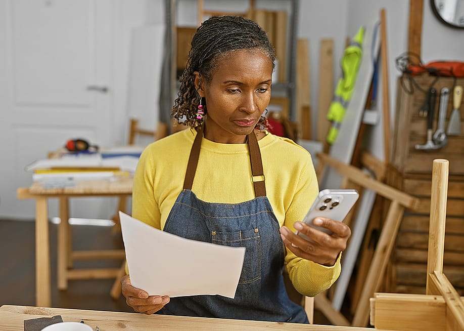 A black female carpentry business owner holds a document and reads information from her smartphone. — Krakenimages.com // Shutterstock