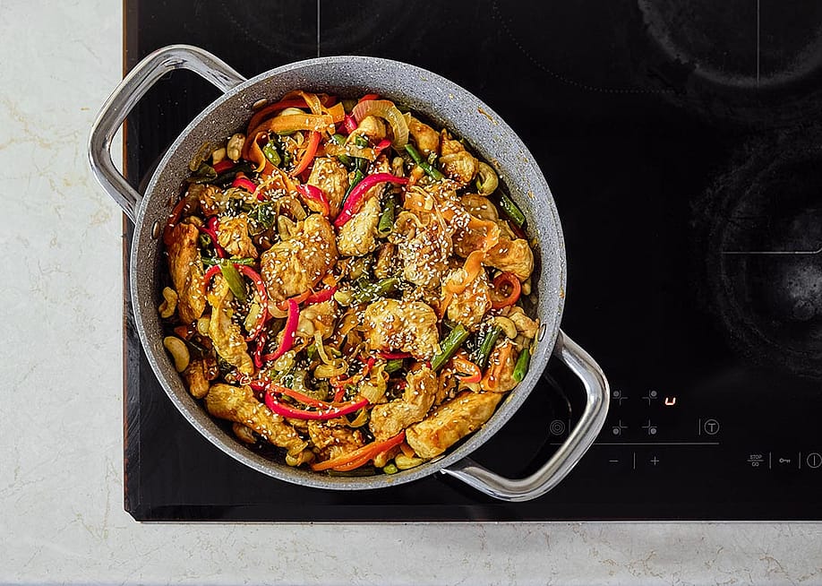 A colorful mix of stir-fried chicken, fresh vegetables, and cashews is sizzling in a pot on an induction cooktop. — Aleksei Isachenko // Shutterstock