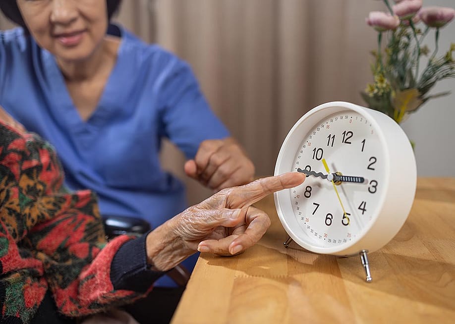 Close up of Asian elderly woman's hand pointing at analog clock with caregiver visible behind her. — Toa55 // Shutterstock