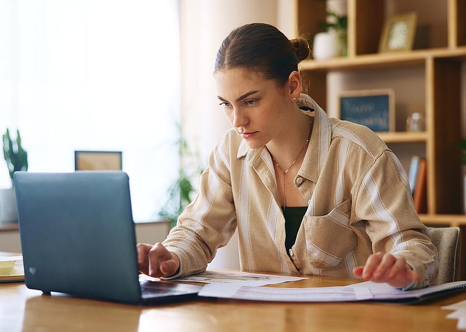 Determined-looking woman looking intently into laptop screen. She is sitting in an office with wooden bookshelves and a window behind her. — PeopleImages // Shutterstock