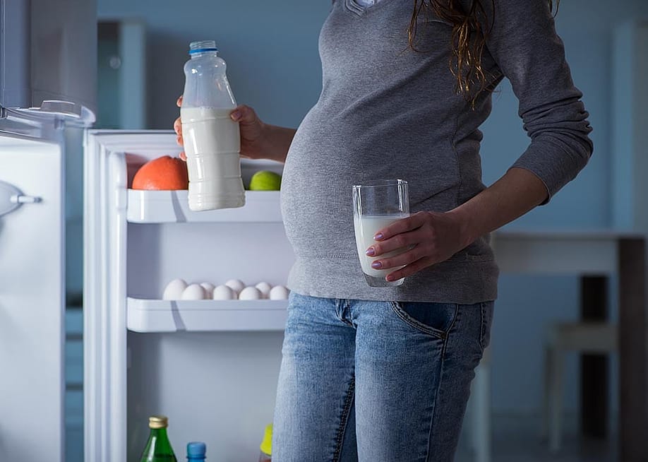 Pregnant woman, whose face is not shown, is illuminated by light of open fridge holding a glass in one hand and bottle of milk in the other. — Elnur // Shutterstock