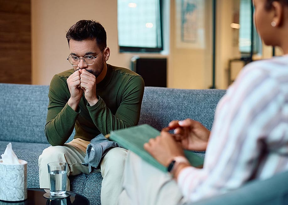 Man in a long sleeve green shirt wearing glasses sits on a blue couch breathing into his hands. A box of tissues is on the table and a therapist's back is in the foreground. — Drazen Zigic // Shutterstock