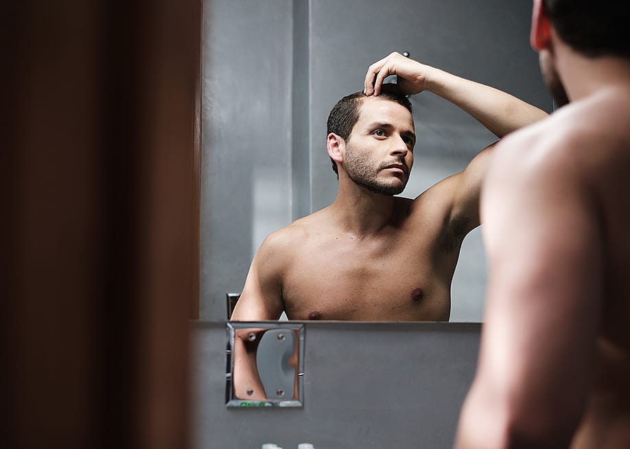 A man looks himself at the mirror inspecting his receding hairline. — engagestock // Shutterstock