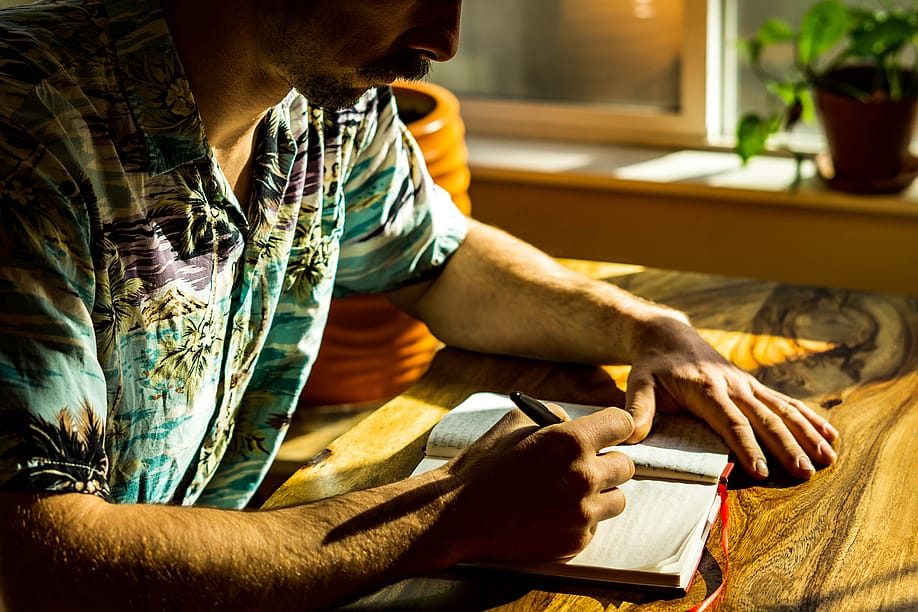 Man wearing a Hawaiian-style shirt sitting next to the window with light coming in, writing in a journal. — Matt Wilenchik // Shutterstock