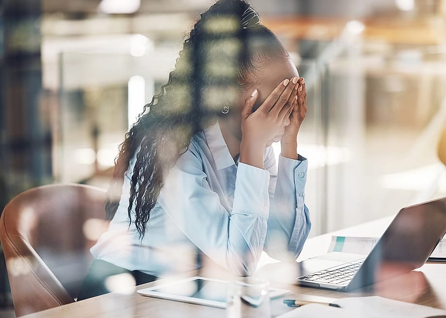 A stressed young black woman with her hands over her face at work. — PeopleImages // Shutterstock