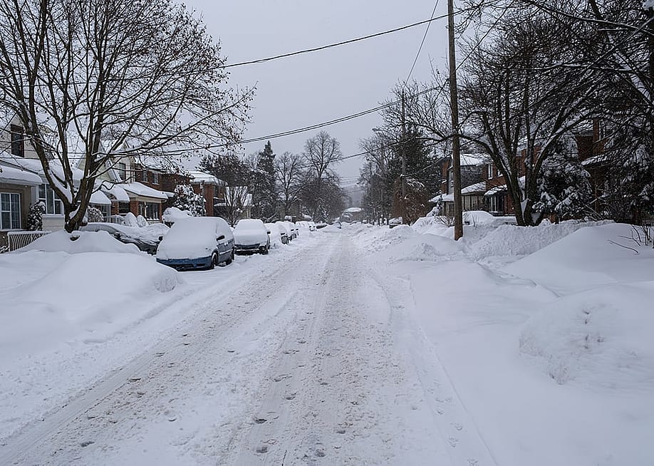 Unplowed snow accumulated on a residential street where rows of cars are parked. — Elena Berd // Shutterstock