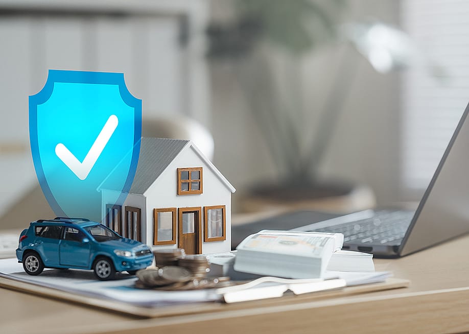 Cash, figures of a house and a car beside a laptop illustrated with a blue safety check graphic as a concept of insurance. — Garun Studios // Shutterstock