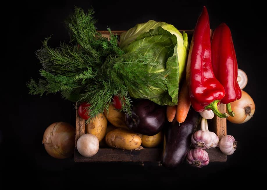 Assortment of beautifully-lit, fresh raw vegetables inlcuding leafy greens and red peppers in a wooden box against black background. — Anna Aybetova // Shutterstock