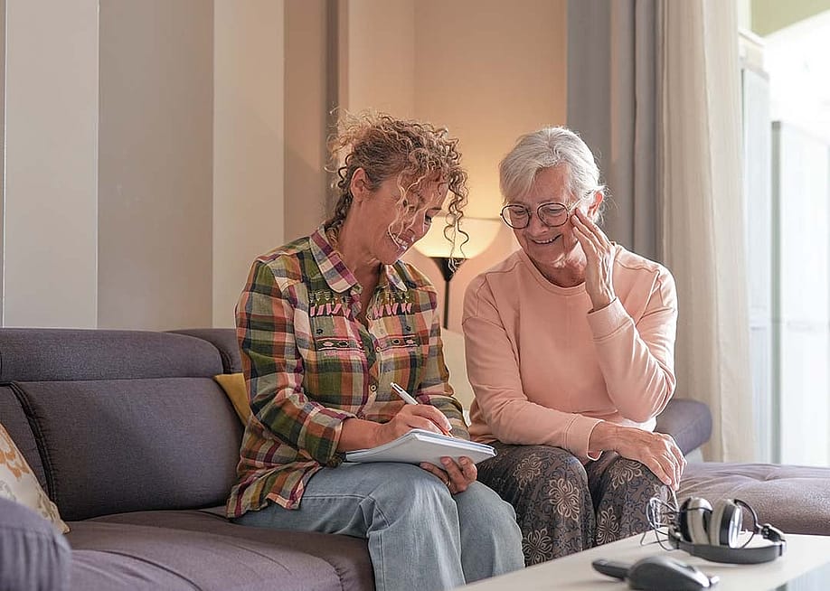 A senior woman and her caregiver sitting together on a sofa at home. — Lucigerma // Shutterstock