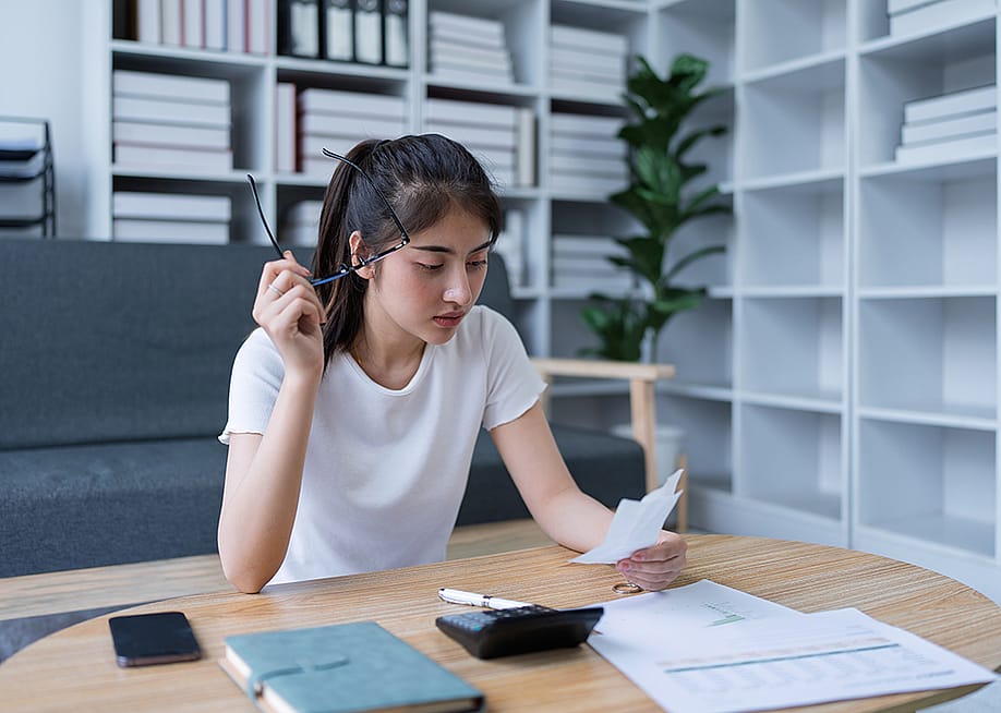 A confused young woman reviewing financial documents at her home office. — Jirapong Manustrong // Shutterstock