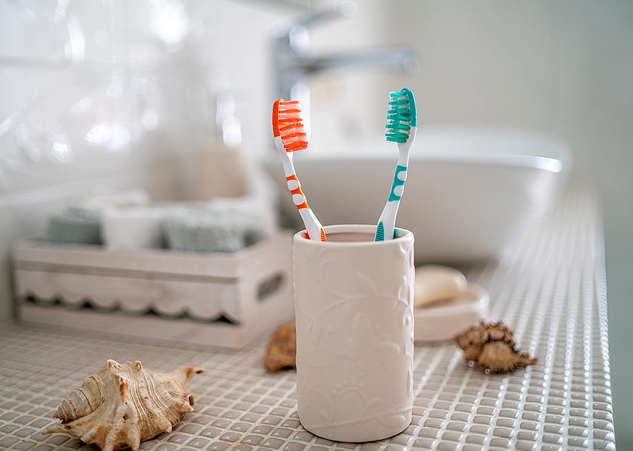 Two colorful toothbrushes in a ceramic glass against the background of a bathroom's washbasin. — Igor Klyakhin // Shutterstock