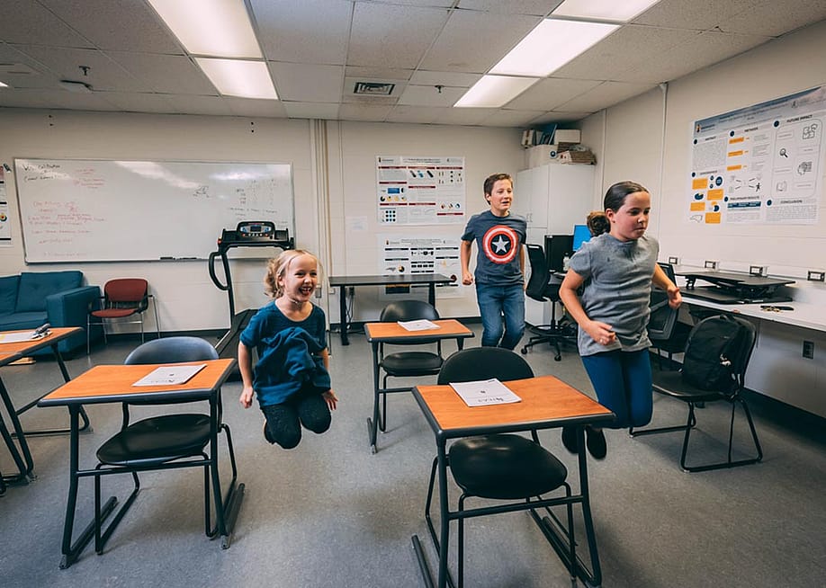 Three elementary-school-age students jumping in the air next to desks in a classroom. — Courtesy of UNC Greensboro University Communications