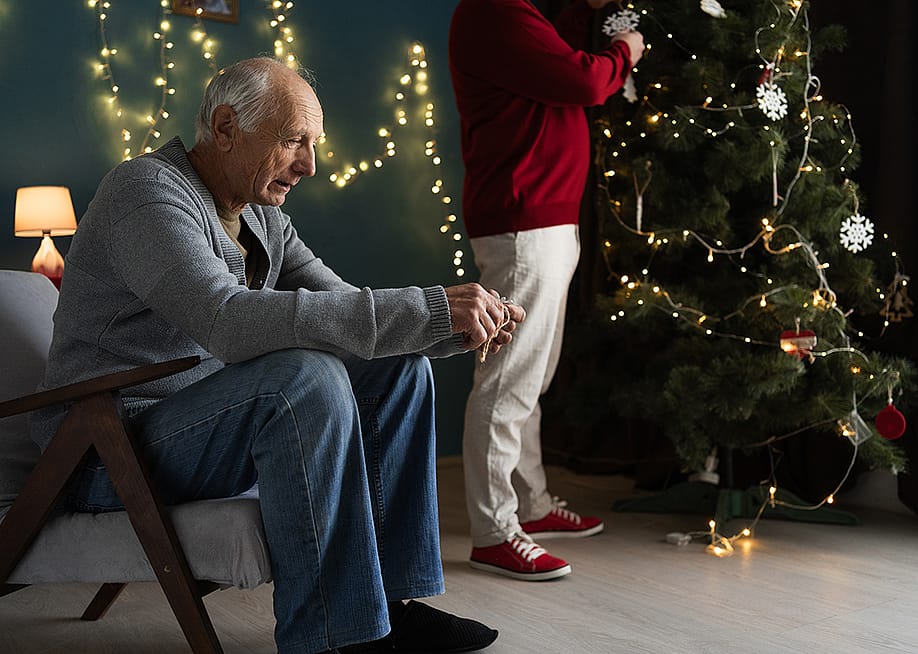A stressed elderly man sitting by their Christmas tree as his partner decorates it. — Lysenko Andrii // Shutterstock