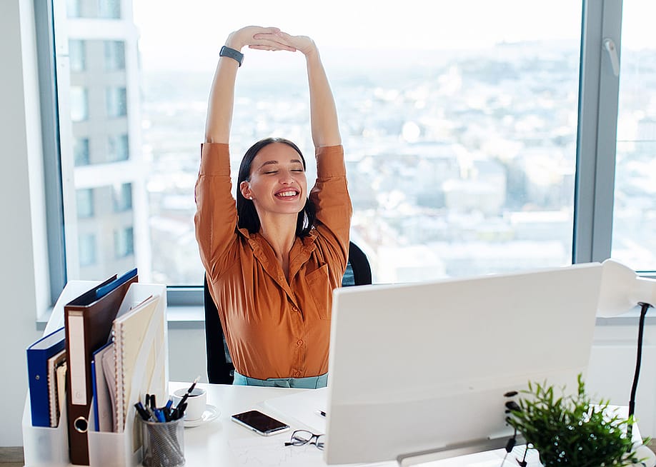 A female manager happily stretching her back and arms while seated at her desk in an office. — fast-stock // Shutterstock
