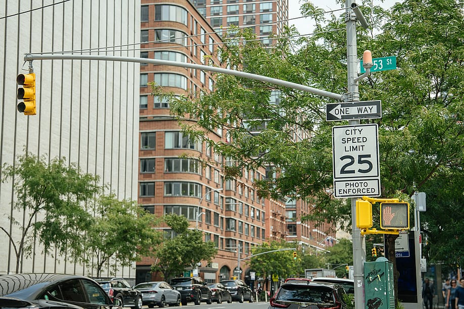 A City Speed Limit 25 sign with a photo enforcement notice stands near a traffic light on a busy urban corner. Modern buildings, cars, and a street sign for 53rd Street are visible in the background. — DenisProduction.com // Shutterstock